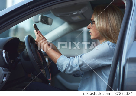 Side view of a concentrated woman or business lady wearing eyeglasses sitting behind steering wheel in her modern car and using smartphone Side view of a concentrated woman or business lady wearing eyeglasses sitting behind steering wheel in her modern car and using smartphone 82853384