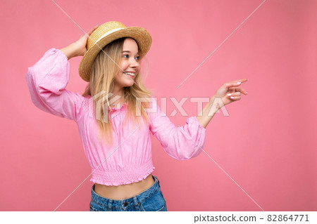 Young beautiful woman in stylish pink summer blouse and trendy straw hat. Positive female shows Young beautiful woman in stylish pink summer blouse and trendy straw hat. Positive female shows 82864771