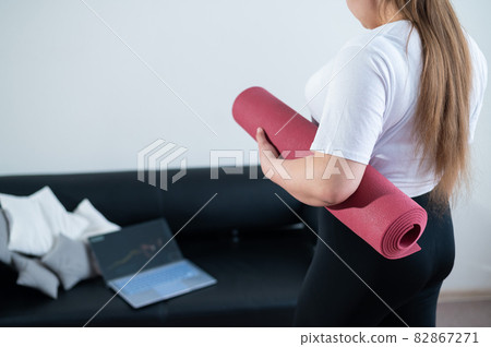 Young chubby woman folds sports mat after online fitness class Young chubby woman folds sports mat after online fitness class 82867271