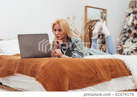 Young woman lying on her bed looking at her laptop screen at Christmas time Young woman lying on her bed looking at her laptop screen at Christmas time 82874179