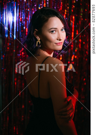 Christmas party look. Elegant woman. Holiday style. Pretty smiling lady purple curly hair posing on shimmering red cascade curtain background blur shadow light. Christmas party look. Elegant woman. Holiday style. Pretty smiling lady purple curly hair posing on shimmering red cascade curtain background blur shadow light. 82877693