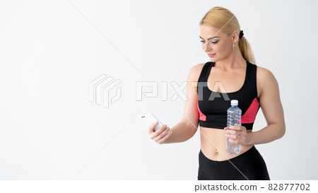 Sportive lifestyle. Athletic woman. Hydration balance. Healthy body. Advertising background. Beautiful lady in sportswear holding bottle of water looking smartphone isolated white copy space. Sportive lifestyle. Athletic woman. Hydration balance. Healthy body. Advertising background. Beautiful lady in sportswear holding bottle of water looking smartphone isolated white copy space. 82877702