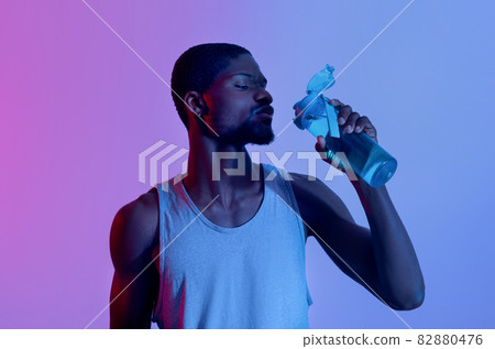 Thirsty young black sportsman drinking water from bottle in neon light 82880476