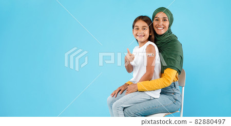 Vaccinated Muslim Mother And Little Daughter Gesturing Thumbs-Up, Blue Background 82880497