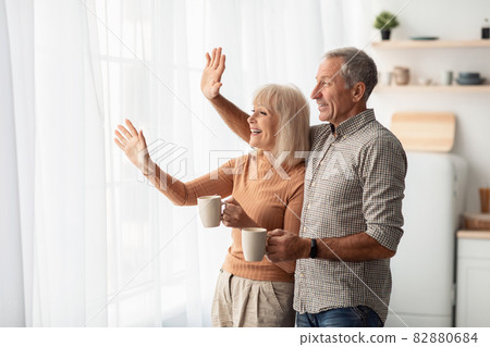 Happy Senior Couple Waving Hello Looking Out Of Window Indoor 82880684