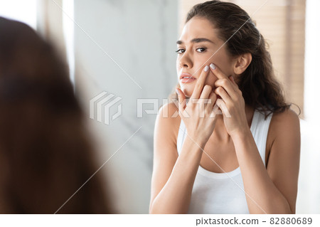 Unhappy Female Squeezing Pimples On Cheek Looking At Mirror Indoor 82880689