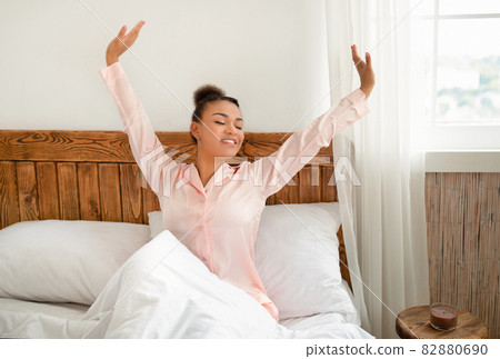 Happy and relaxed after good night sleep. Young african american lady in pink pajamas stretching after waking up 82880690