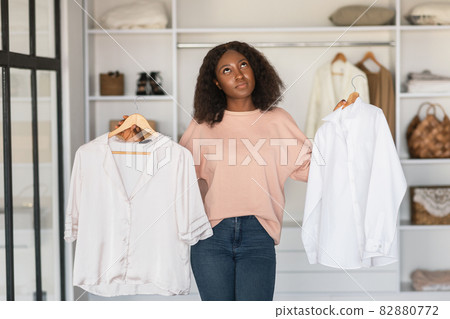 Pensive Black Woman Thinking Holding Two White Shirts At Home 82880772