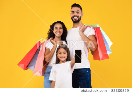 Middle-Eastern Family Showing Smartphone Posing With Shopping Bags, Yellow Background 82880839