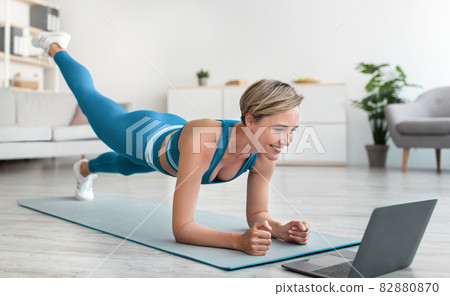 Happy woman doing plank with leg raise using laptop 82880870
