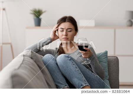 Sad indifferent caucasian young lady sits on sofa with glass of wine and suffering from headache in home Sad indifferent caucasian young lady sits on sofa with glass of wine and suffering from headache in home 82881064