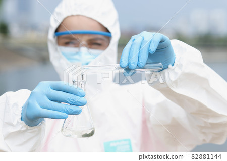 Scientist in protective suit with glasses pours water from test tube into flask Scientist in protective suit with glasses pours water from test tube into flask 82881144