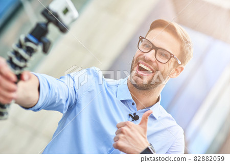 Young happy man, male blogger wearing blue shirt holding a gimbal with smartphone and recording video for his blog outdoors, smiling at camera and showing thumb up 82882059