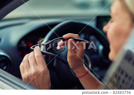 Side view of a woman holding eyeglasses and smiling while sitting behind steering wheel of her modern car, driving through the city Side view of a woman holding eyeglasses and smiling while sitting behind steering wheel of her modern car, driving through the city 82883756