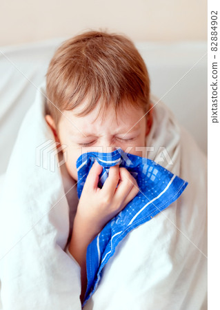 the boy blows his nose in a handkerchief, wrapped in a blanket. sick child. a cold with a runny nose in a boy the boy blows his nose in a handkerchief, wrapped in a blanket. sick child. a cold with a runny nose in a boy 82884902