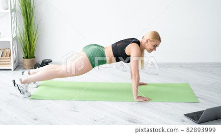 Fitness lifestyle. Athletic woman. Home training. Online sport. Happy fit lady in sportswear doing plank workout on yoga mat looking laptop light room interior. Fitness lifestyle. Athletic woman. Home training. Online sport. Happy fit lady in sportswear doing plank workout on yoga mat looking laptop light room interior. 82893999