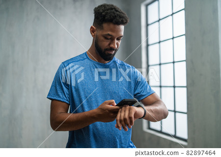 Happy young African American sportsman standing indoors at gym, using smartphone and smartwatch. Happy young African American sportsman standing indoors at gym, using smartphone and smartwatch. 82897464