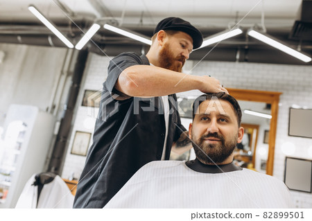 Two handsome men at barbershop. Stylish red-bearded barber and client during haircut process. Beauty, healthcare and male cosmetics concept. 82899501