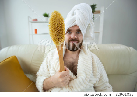 Portrait of funny guy wear turban towel holding in hands massage brush while sits on sofa. Male skin care and spa concept. 82901853