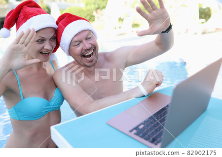 Young man and woman in red santa hats waving at laptop screen on side of pool Young man and woman in red santa hats waving at laptop screen on side of pool 82902175