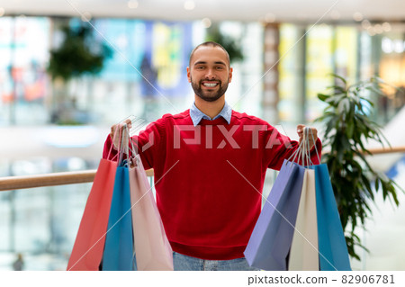 Portrait of happy young man holding colorful shopper bags in outstretched hands at big supermarket 82906781