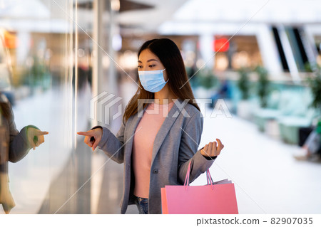 Young Asian woman in face mask holding gift bags, shopping for new clothes at huge supermarket 82907035