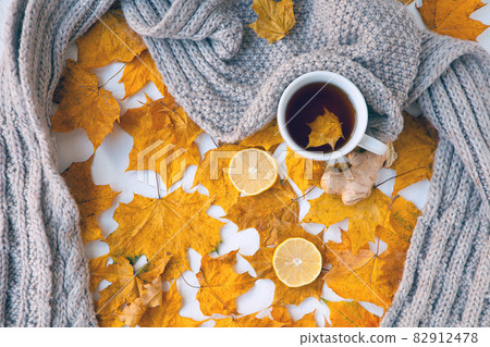 Autumn still life with white cup of tea, warming grey scarf and colorful yellow leaves with ginger and lemon. Selective focus on mug, shallow. Autumn still life with white cup of tea, warming grey scarf and colorful yellow leaves with ginger and lemon. Selective focus on mug, shallow. 82912478