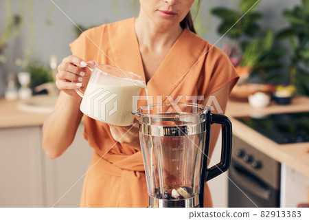 Woman in orange dress pouring fresh milk into electric blender Woman in orange dress pouring fresh milk into electric blender 82913383