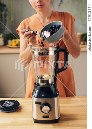 Young woman putting fresh blueberries into electric blender 82913384