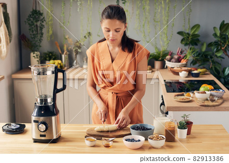 Brunette woman cutting banana by kitchen table 82913386
