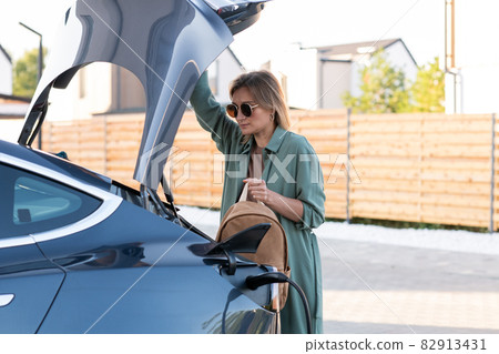 Young blond woman taking backpack out of trunk while car being charged 82913431