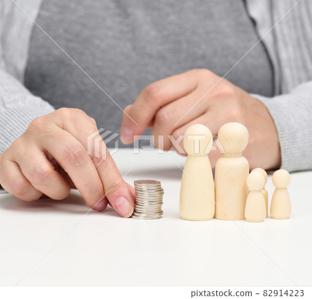 hand puts coins in a pile and wooden figurines of a family on a white table. Economy and investment concept, budget planning. Receiving subsidies from the state hand puts coins in a pile and wooden figurines of a family on a white table. Economy and investment concept, budget planning. Receiving subsidies from the state 82914223