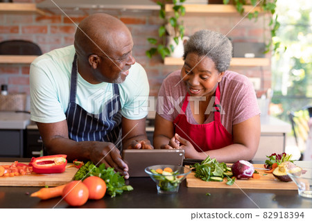Happy african american senior couple cooking together in kitchen, using tablet 82918394
