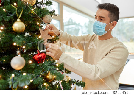 man in mask decorating christmas tree at home 82924593