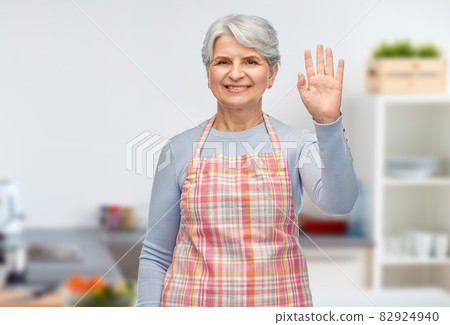 smiling senior woman in kitchen apron waving hand 82924940
