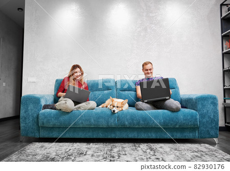Two smiling people sitting on different ends of blue sofa. Woman invisibly looking into her husband's laptop. Pet dog in glasses is bored, waiting for person to pay attention or play with him Two smiling people sitting on different ends of blue sofa. Woman invisibly looking into her husband's laptop. Pet dog in glasses is bored, waiting for person to pay attention or play with him 82930176
