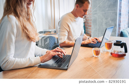 Close up of young woman and man typing on laptop keyboard while spending time together at home. Married couple sitting at wooden table with cups of tea and using modern notebooks. 82930191