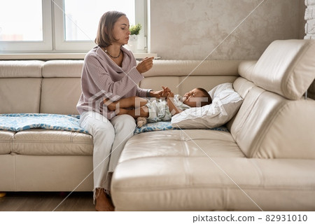 Worried young mother sitting on sofa beside her sick son with high fever. Mom measures temperature 82931010
