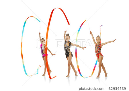 Three-ribbon event. Portrait of little beautiful rhythmic gymnastics artists in group sport event isolated on white studio background. Three-ribbon event. Portrait of little beautiful rhythmic gymnastics artists in group sport event isolated on white studio background. 82934589