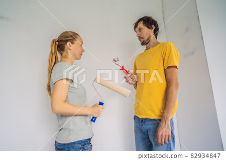 A man and a woman with rollers for painting the walls. A man with a very small bolster, symbolizing a small penis. renovation diy paint couple in new home painting wall 82934847
