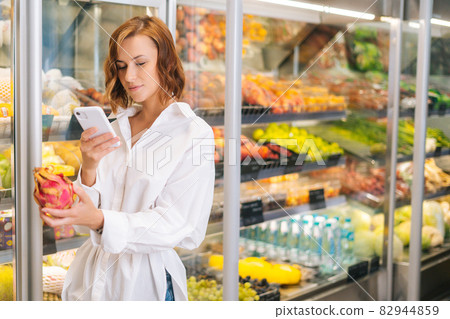 Medium shot of pretty young woman holding exotic fruit and looking for it name on mobile phone. Medium shot of pretty young woman holding exotic fruit and looking for it name on mobile phone. 82944859