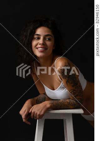 Cropped portrait of young attractive woman in white inner wear posing on chair isolated over dark background Cropped portrait of young attractive woman in white inner wear posing on chair isolated over dark background 82946150