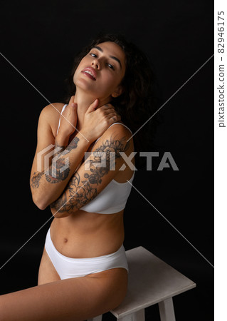 Cropped studio portrait of beautiful young curly woman posing in white cotton underwear, sitting on chair isolated over dark background Cropped studio portrait of beautiful young curly woman posing in white cotton underwear, sitting on chair isolated over dark background 82946175