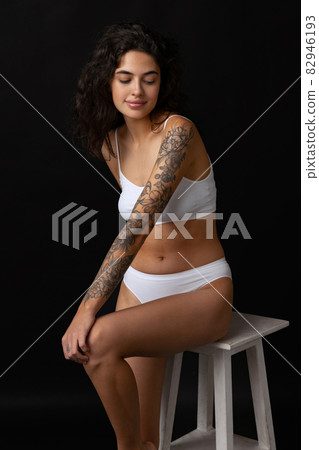 Cropped portrait of beautiful girl in white cotton underwear posing on chair isolated over dark background. Body positivity concept 82946193