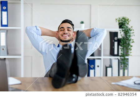 Happy millennial Arab man relaxing after successful work, putting feet on table at modern office 82946354