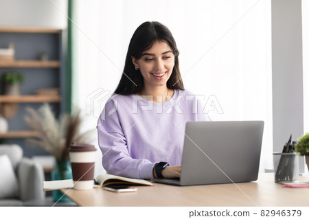 Cheerful millennial lady sitting at desk using laptop 82946379