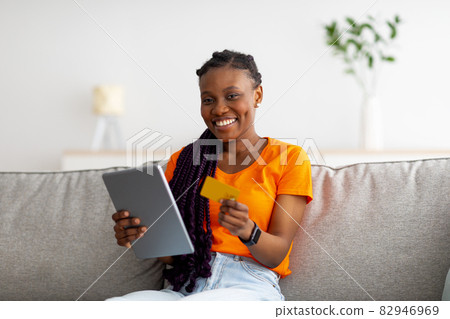 Positive black lady shopping online at home, using tablet and credit card, sitting on couch and smiling at camera Positive black lady shopping online at home, using tablet and credit card, sitting on couch and smiling at camera 82946969