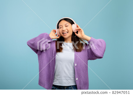 Happy asian lady listening to music in wireless headphones, closing eyes and relaxing on blue studio background 82947051