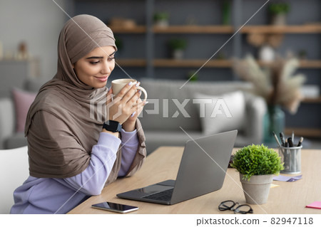 Muslim lady watching video on pc, holding cup drinking coffee Muslim lady watching video on pc, holding cup drinking coffee 82947118