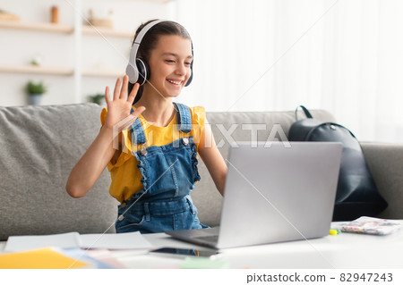 Cheerful girl sitting at table, using laptop, waving to webcam 82947243